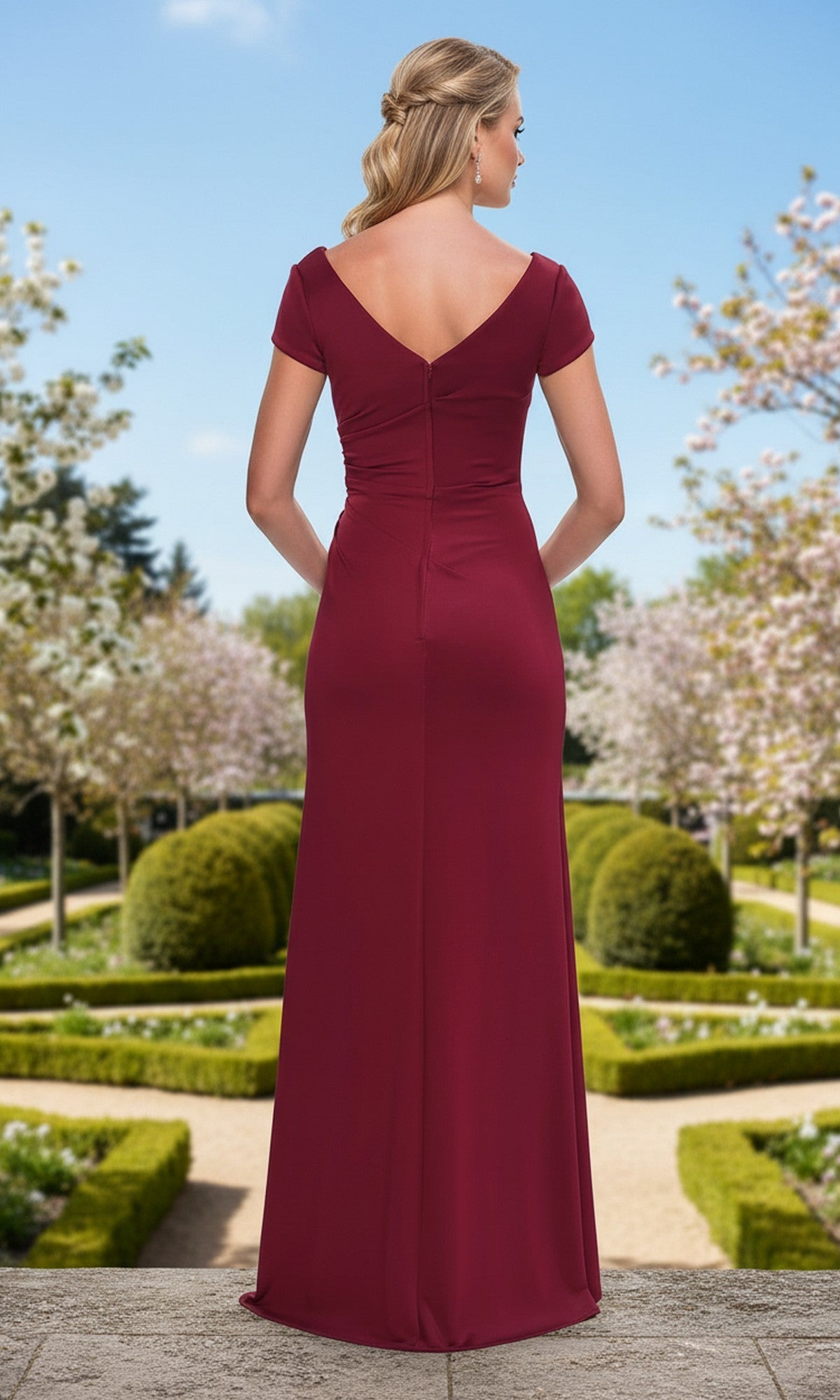 A woman stands with her back to the camera in a formal garden, wearing the Long Prom Dress: La Femme 33616, a floor-length column dress with short sleeves.